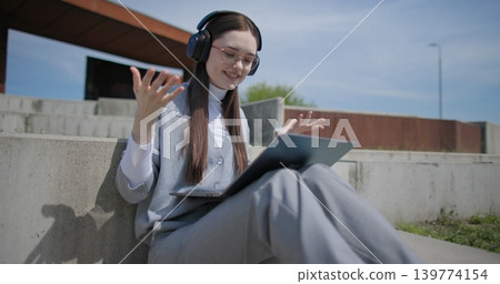 A hand-held shot of a young female student in casual clothes, glasses, and headphones making a video call on a laptop while sitting on a concrete bench in an urban park. A hand-held shot of a young female student in casual clothes, glasses, and headphones making a video call on a laptop while sitting on a concrete bench in an urban park. 139774154