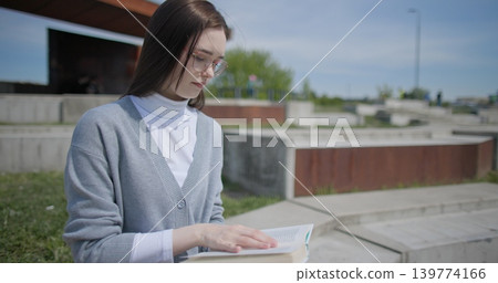 A hand-held shot of a young female student in casual clothes and glasses reading a book while flipping through pages while sitting in an urban park. 139774166