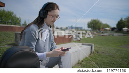 A hand-held shot of a young female student in casual clothes, wearing glasses and headphones, sitting on a concrete in an urban park on a sunny summer day and listening to music from a mobile phone 139774168