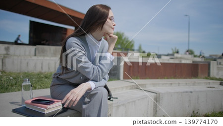 A handheld shot of a young female student in casual clothes sitting on a concrete bench in an urban park, enjoying a sunny summer day. A handheld shot of a young female student in casual clothes sitting on a concrete bench in an urban park, enjoying a sunny summer day. 139774170