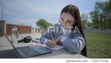 A hand-held shot of a young female student in casual clothes and glasses using a laptop and taking notes with a pen in a notebook in an urban park. A hand-held shot of a young female student in casual clothes and glasses using a laptop and taking notes with a pen in a notebook in an urban park. 139774188
