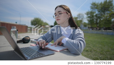 A hand-held shot of a young female student in casual clothes and glasses using a laptop and taking notes with a pen in a notebook in an urban park. 139774199