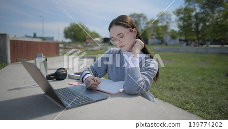 A hand-held shot of a young female student in casual clothes and glasses using a laptop and taking notes with a pen in a notebook in an urban park. A hand-held shot of a young female student in casual clothes and glasses using a laptop and taking notes with a pen in a notebook in an urban park. 139774202