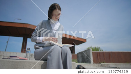 A hand-held shot of a young female student in casual clothes and glasses reading a book while flipping through pages while sitting in an urban park. A hand-held shot of a young female student in casual clothes and glasses reading a book while flipping through pages while sitting in an urban park. 139774219