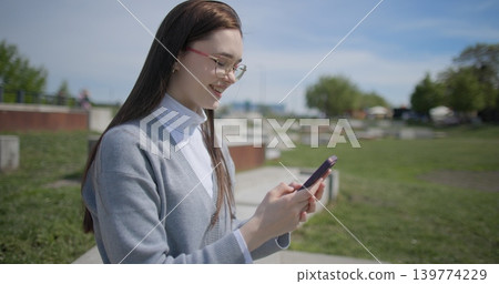 A hand-held shot of a young female student in casual clothes wearing glasses sitting on a concrete bench in an urban park on a sunny summer day using a mobile phone. A hand-held shot of a young female student in casual clothes wearing glasses sitting on a concrete bench in an urban park on a sunny summer day using a mobile phone. 139774229