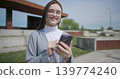 A hand-held shot of a young female student in casual clothes wearing glasses sitting on a concrete bench in an urban park on a sunny summer day using a mobile phone. 139774240