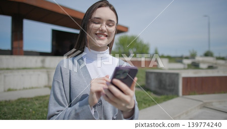 A hand-held shot of a young female student in casual clothes wearing glasses sitting on a concrete bench in an urban park on a sunny summer day using a mobile phone. 139774240