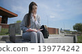 A hand-held portrait of a young female student in casual clothes wearing glasses, sitting on a concrete bench in an urban park, leafing through a notebook and taking notes with a pen. 139774242