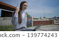 A hand-held shot of a young female student in casual clothes and glasses conducting a video call or conference on a laptop while sitting in an urban park 139774243