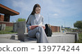 A hand-held portrait of a young female student in casual clothes wearing glasses, sitting on a concrete bench in an urban park, leafing through a notebook and taking notes with a pen. 139774244