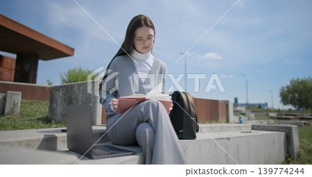 A hand-held portrait of a young female student in casual clothes wearing glasses, sitting on a concrete bench in an urban park, leafing through a notebook and taking notes with a pen. A hand-held portrait of a young female student in casual clothes wearing glasses, sitting on a concrete bench in an urban park, leafing through a notebook and taking notes with a pen. 139774244