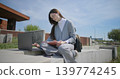 A hand-held shot of a young female student in casual clothes and glasses using a laptop and taking notes with a pen in a notebook in an urban park. 139774245