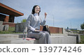 A hand-held shot of a young female student in casual clothes, glasses, and headphones working and typing on a laptop while sitting on a concrete bench in an urban park. 139774246