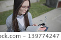 A hand-held shot of a young female student in casual clothes wearing glasses sitting on a concrete bench in an urban park on a sunny summer day and reading a book, flipping through the pages. 139774247