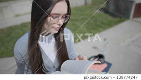 A hand-held shot of a young female student in casual clothes wearing glasses sitting on a concrete bench in an urban park on a sunny summer day and reading a book, flipping through the pages. A hand-held shot of a young female student in casual clothes wearing glasses sitting on a concrete bench in an urban park on a sunny summer day and reading a book, flipping through the pages. 139774247