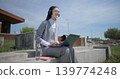 A hand-held shot of a young female student in casual clothes, glasses, and headphones working and typing on a laptop while sitting on a concrete bench in an urban park. 139774248