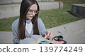 A hand-held shot of a young female student in casual clothes wearing glasses sitting on a concrete bench in an urban park on a sunny summer day and reading a book, flipping through the pages. 139774249
