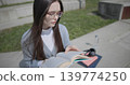 A hand-held shot of a young female student in casual clothes wearing glasses sitting on a concrete bench in an urban park on a sunny summer day and reading a book, flipping through the pages. 139774250