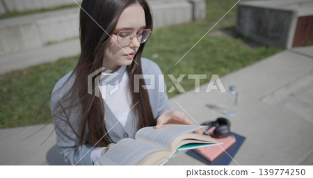 A hand-held shot of a young female student in casual clothes wearing glasses sitting on a concrete bench in an urban park on a sunny summer day and reading a book, flipping through the pages. 139774250