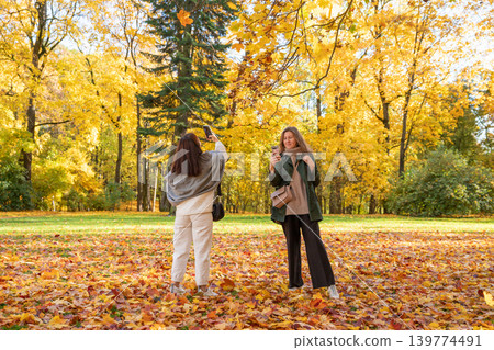 Woman taking photo of friend with smartphone in autumn park. Two women enjoying fall season outdoors. Golden leaf foliage in forest during weekend leisure. Friends spending time together. Woman taking photo of friend with smartphone in autumn park. Two women enjoying fall season outdoors. Golden leaf foliage in forest during weekend leisure. Friends spending time together. 139774491