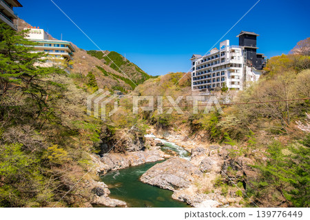 Nikko City, Tochigi Prefecture: A view of the Kinugawa Gorge and hot spring town from Takimi Bridge, under a wide blue sky and surrounded by lush greenery. 139776449