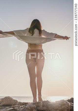 Woman beach sunset young woman in bikini and white shirt stands on coastal rocks enjoying freedom at the sea Woman beach sunset young woman in bikini and white shirt stands on coastal rocks enjoying freedom at the sea 139777340