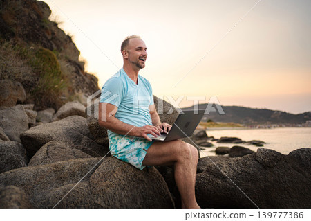 Mid adult man happily remote working on beach during sunset Mid adult man happily remote working on beach during sunset 139777386