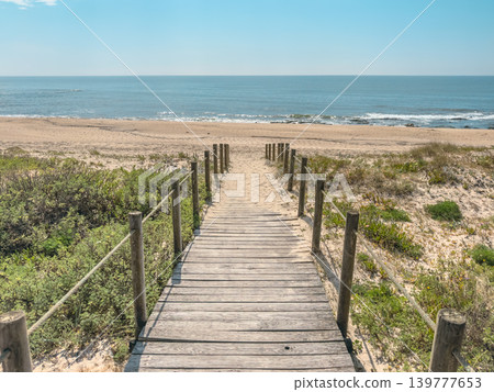 Wide perspective of a rustic wooden boardwalk leading through coastal dunes to the sandy shore and blue horizon of Sao Felix da Marinha beach in Porto Portugal Wide perspective of a rustic wooden boardwalk leading through coastal dunes to the sandy shore and blue horizon of Sao Felix da Marinha beach in Porto Portugal 139777653