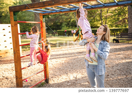 Smiling mom helps her cute daughter climb the playground Smiling mom helps her cute daughter climb the playground 139777697