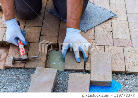 Worker uses hammer to lay bricks on path while applying cement in residential area under patio renovation Worker uses hammer to lay bricks on path while applying cement in residential area under patio renovation 139778377