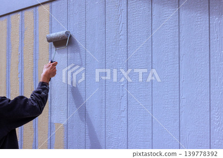 Painter uses paint roller to apply light gray paint on vertical wooden siding wall of building. Painter uses paint roller to apply light gray paint on vertical wooden siding wall of building. 139778392