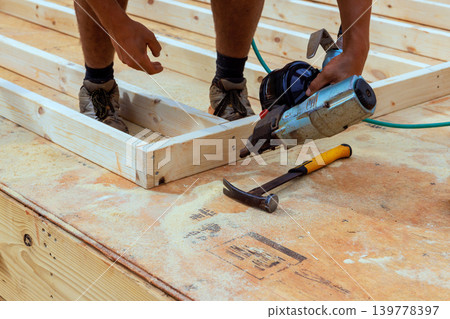Worker uses nail gun to attach wooden beams while standing on wooden floor at construction site. 139778397