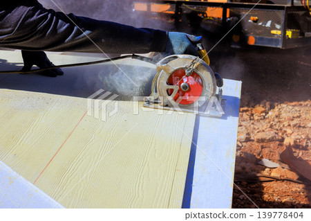 Worker cuts piece of wood with circular hand saw while standing at construction site Worker cuts piece of wood with circular hand saw while standing at construction site 139778404