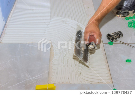 Worker applies tile adhesive with trowel on floor in bathroom renovation project. Worker applies tile adhesive with trowel on floor in bathroom renovation project. 139778427