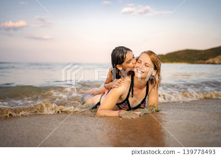 Mother and daughter smiling, lying in waves on a sandy beach Mother and daughter smiling, lying in waves on a sandy beach 139778493