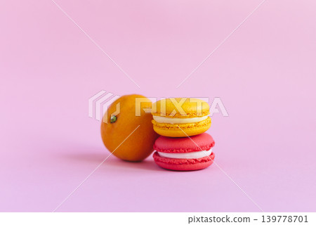 Two tasty french macarons and tangerine on pink background. Pink and yellow macarons. Two tasty french macarons and tangerine on pink background. Pink and yellow macarons. 139778701