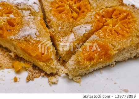 Slices of cake show orange fruit pieces on top, resting on a white plate. The cake is partially cut, and crumbs surround it on the kitchen surface Slices of cake show orange fruit pieces on top, resting on a white plate. The cake is partially cut, and crumbs surround it on the kitchen surface 139778899