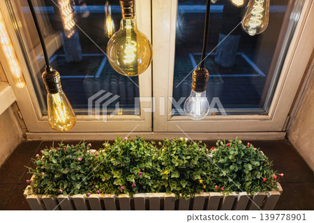 Hanging light bulbs shine above green plants in a windowbox. The scene shows a balcony with bright lights illuminating the night Hanging light bulbs shine above green plants in a windowbox. The scene shows a balcony with bright lights illuminating the night 139778901
