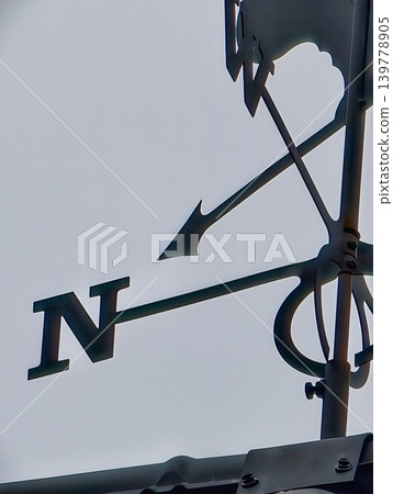 A wind vane shows the direction of the wind with the arrow pointing north against a clear sky. This takes place on a rooftop in the late afternoon 139778905