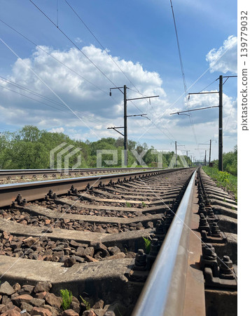 Railway tracks with electric power lines stretching into distance under blue cloudy sky Railway tracks with electric power lines stretching into distance under blue cloudy sky 139779032