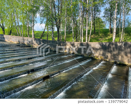 Cascading water flow in concrete canal surrounded by green trees and hydraulic infrastructure Cascading water flow in concrete canal surrounded by green trees and hydraulic infrastructure 139779042