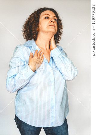 Middle-aged woman in blue shirt touching her neck and chest showing discomfort and concern Middle-aged woman in blue shirt touching her neck and chest showing discomfort and concern 139779128