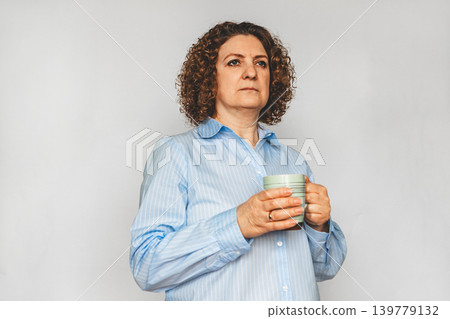 Thoughtful middle-aged woman holding a mug and gazing into the distance in a blue shirt Thoughtful middle-aged woman holding a mug and gazing into the distance in a blue shirt 139779132