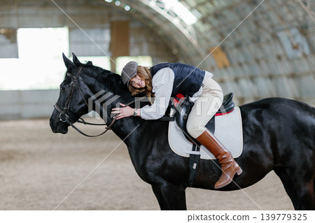 Young woman embracing black horse in indoor arena Young woman embracing black horse in indoor arena 139779325