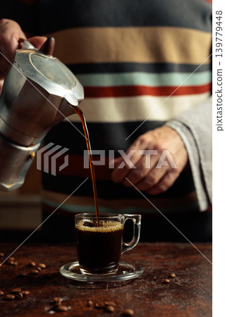 A man in a striped sweater is poured coffee from a geyser coffee maker into a glass cup. A man in a striped sweater is poured coffee from a geyser coffee maker into a glass cup. 139779448