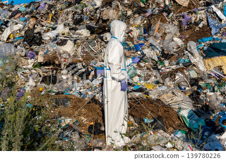 A man in a protective suit stands near a large pile of garbage in a city dump site during daytime hours A man in a protective suit stands near a large pile of garbage in a city dump site during daytime hours 139780226