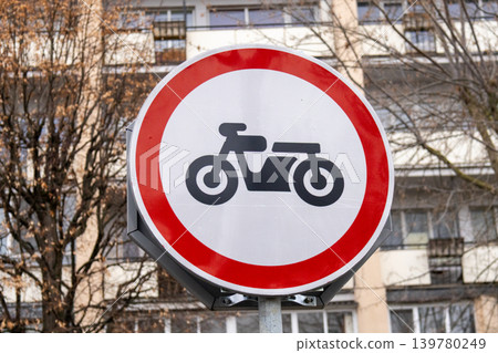 A sign banning motorcycles stands on a post in front of buildings during the day indicating restricted vehicle access A sign banning motorcycles stands on a post in front of buildings during the day indicating restricted vehicle access 139780249