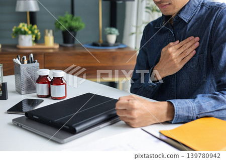 Businessman Experiencing Sudden Chest Pain at Work, Clutching Chest Next to Medicine Bottles on Desk. Businessman Experiencing Sudden Chest Pain at Work, Clutching Chest Next to Medicine Bottles on Desk. 139780942