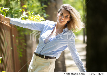Woman leans on a gate during a sunny day in a park with trees around and a path in the background Woman leans on a gate during a sunny day in a park with trees around and a path in the background 139781698