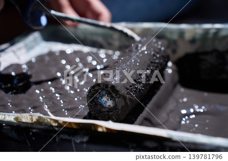 Close up of paint roller resting in tray with dark paint and hand in background Close up of paint roller resting in tray with dark paint and hand in background 139781796